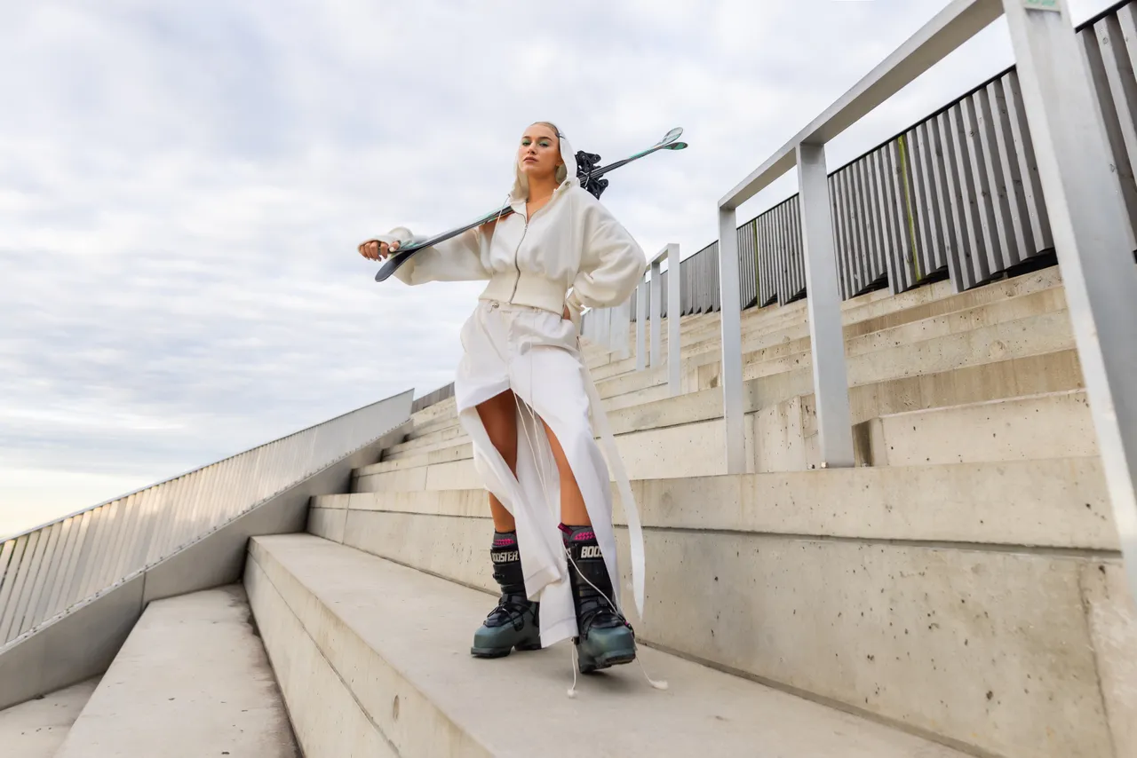 Kelly Sildaru in ski boots holding a ski while standing on concrete stairs.