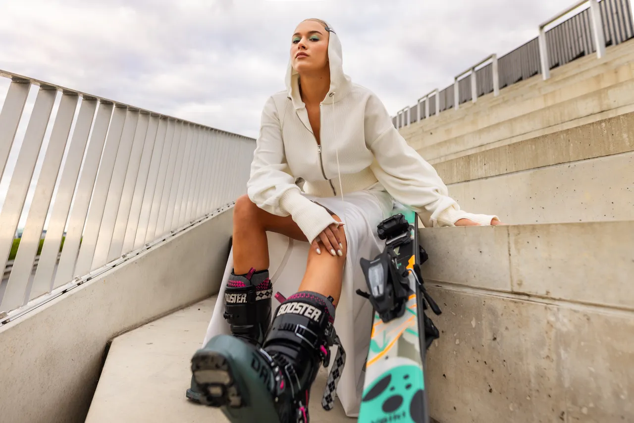 Kelly Sildaru in ski boots holding a ski while standing on concrete stairs.