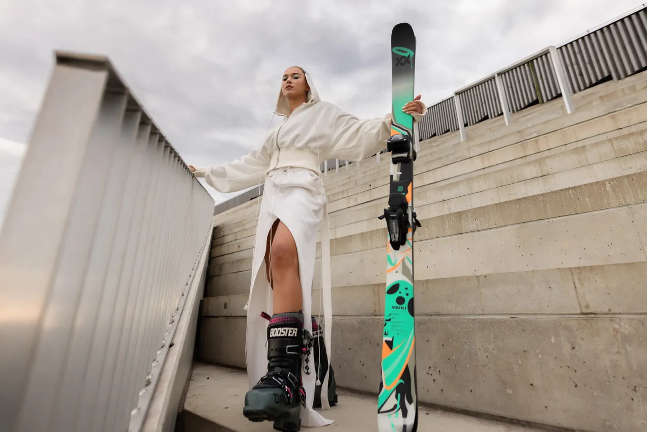 Kelly Sildaru in ski boots holding a ski while standing on concrete stairs.