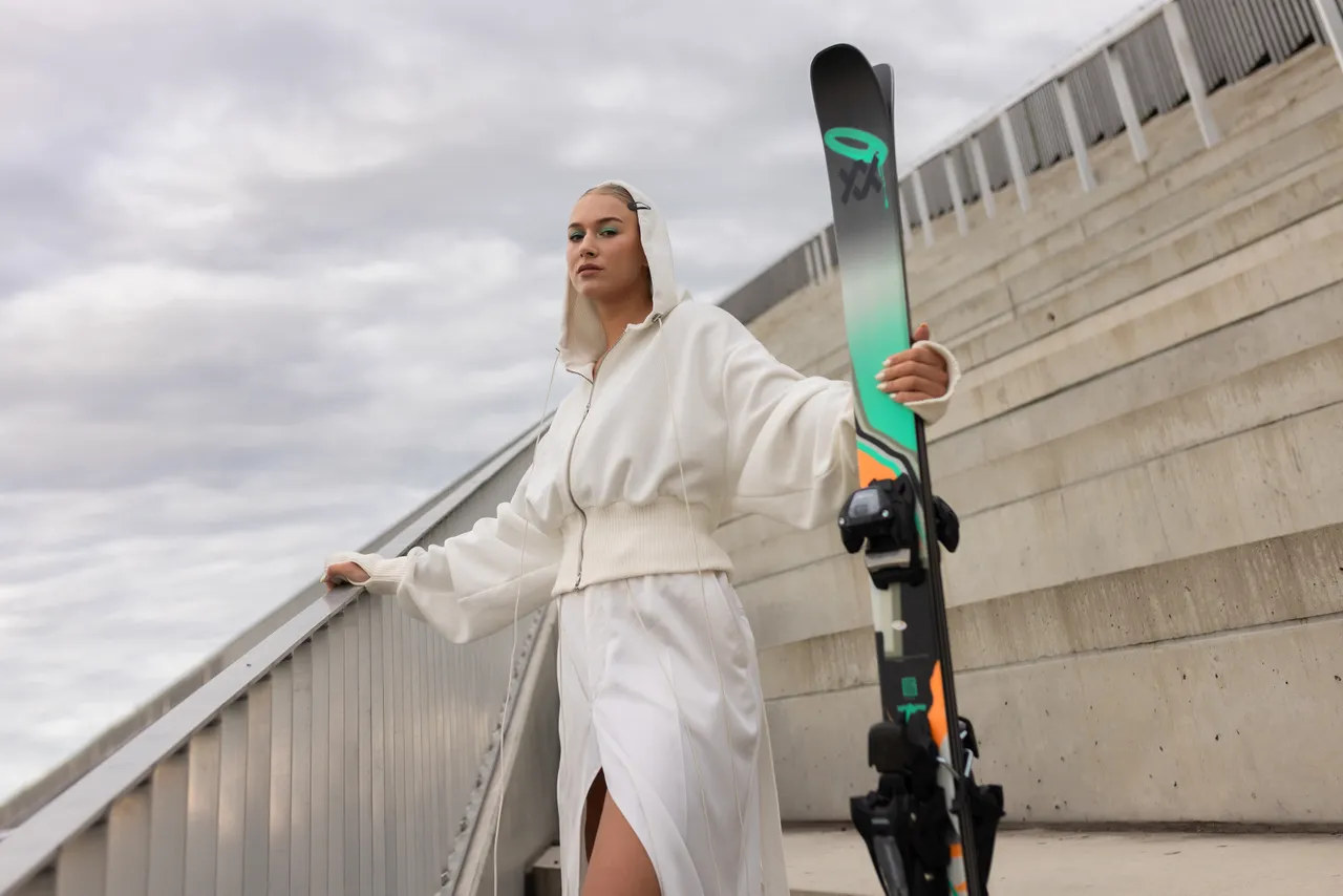 Kelly Sildaru in ski boots holding a ski while standing on concrete stairs.