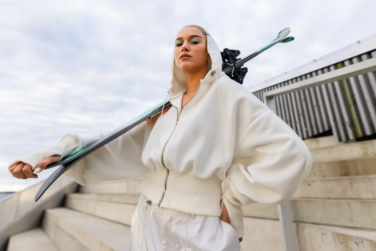 Kelly Sildaru in ski boots holding a ski while standing on concrete stairs.