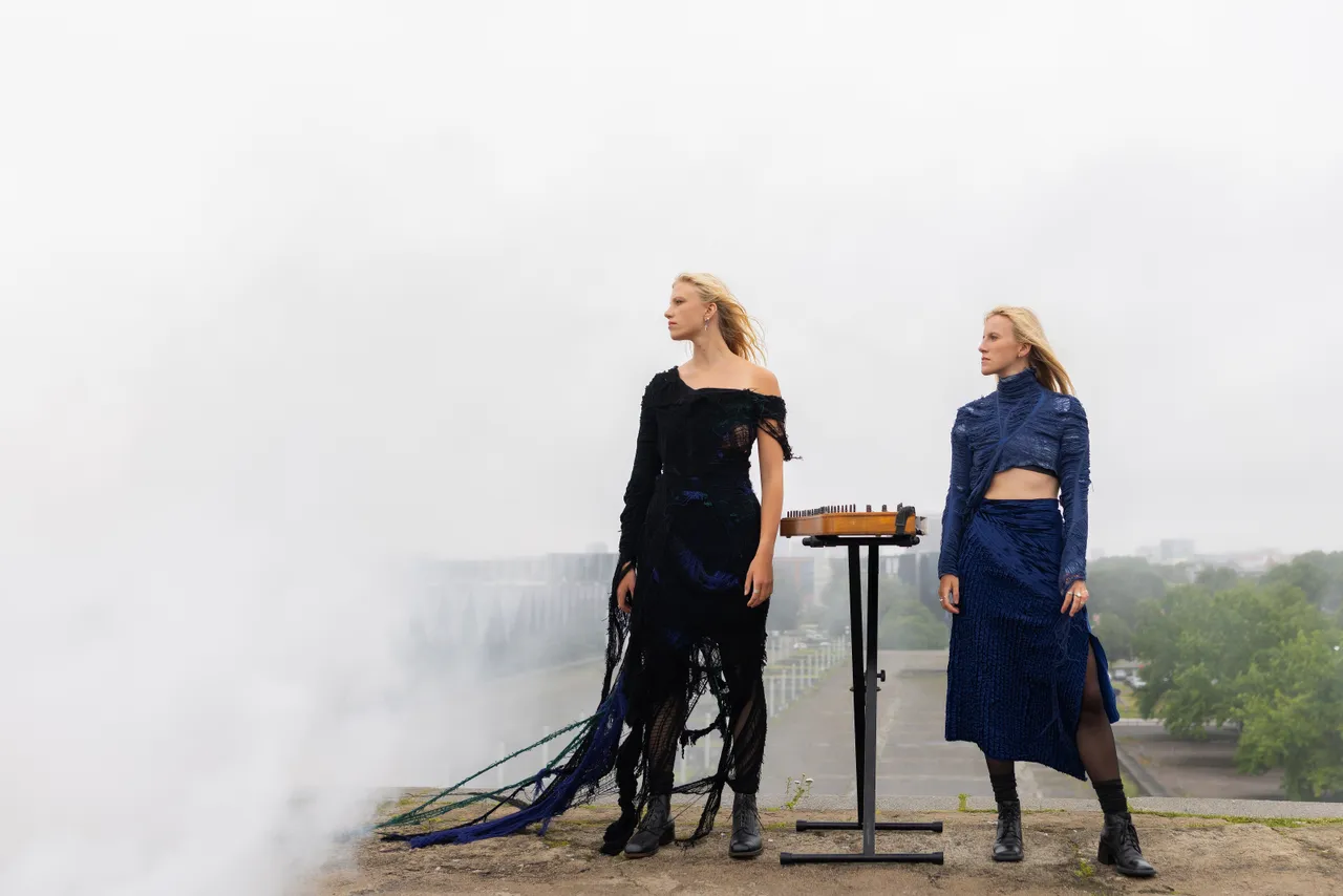 Two women from Duo Ruut, Katariina Kivi and Ann-Lisett Rebane, playing kannel outdoors in a misty urban setting.
