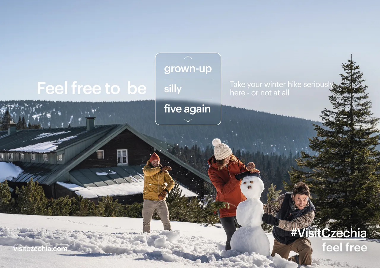 Three people build a snowman in the snow beside a mountain lodge.