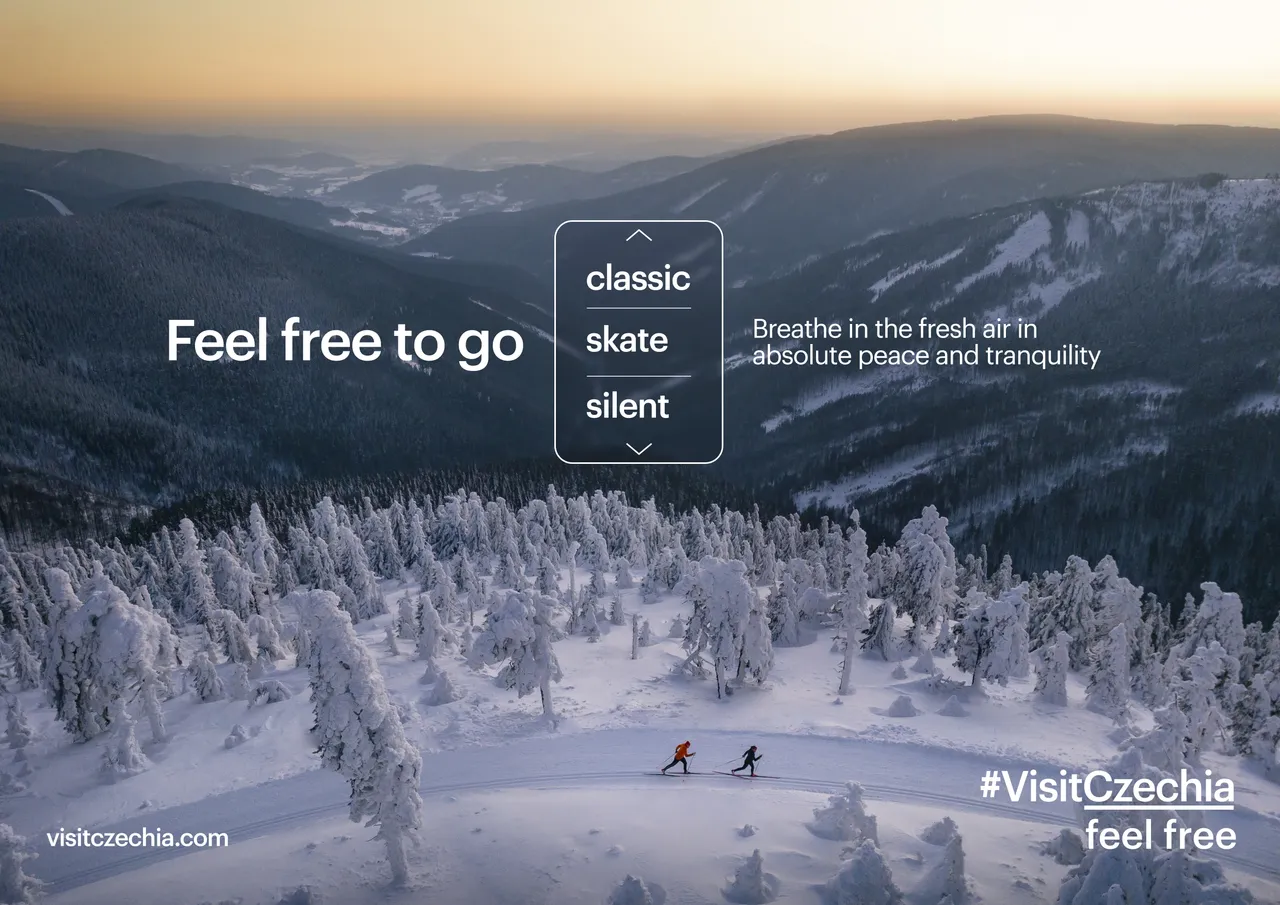 Two skiers cross a snowy clearing surrounded by frosted trees and mountain peaks at sunset.